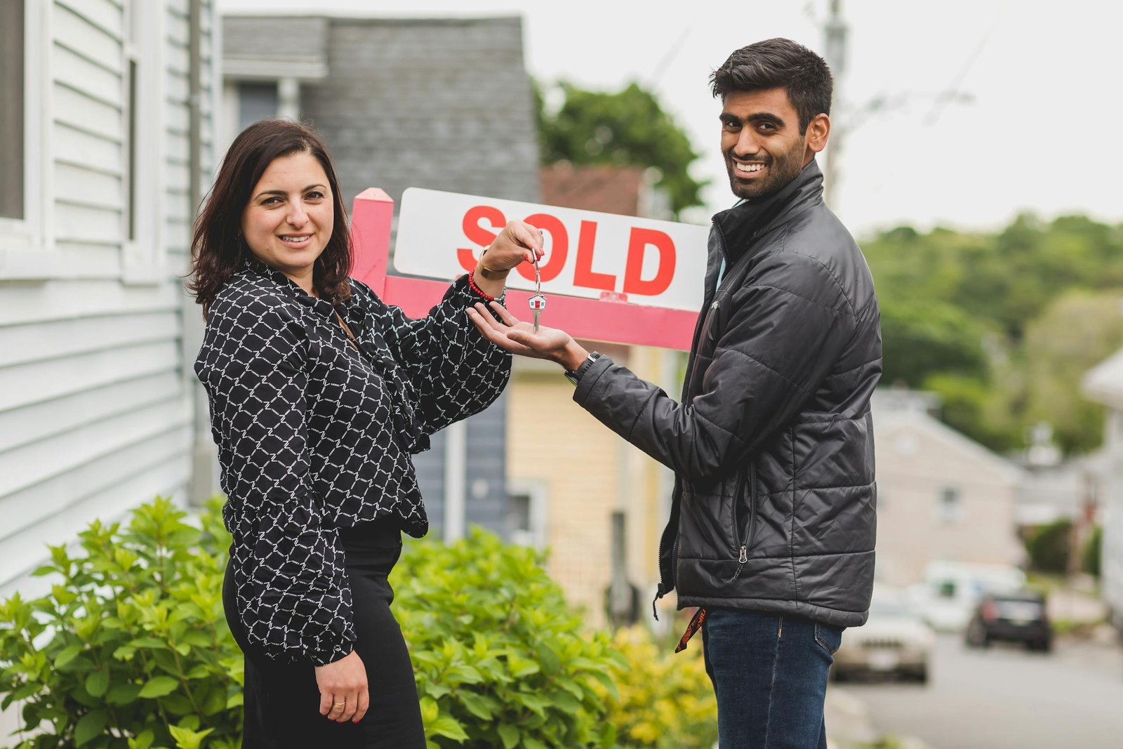 A joyful couple celebrates buying their new home with a key in front of a sold sign.